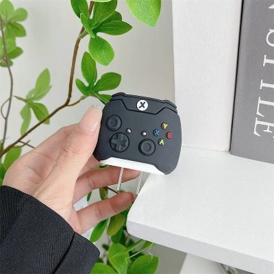 A black VR gaming controller resembling an Xbox gamepad, designed for immersive virtual reality experiences, is held in a person's hand near a desk and greenery.