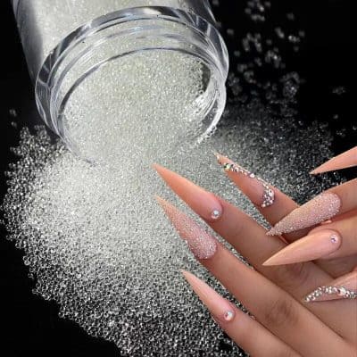 Close-up of a small jar spilling rhinestones onto a black surface, with a hand showcasing long, decorated acrylic nails adorned with rhinestones for a glamorous look.