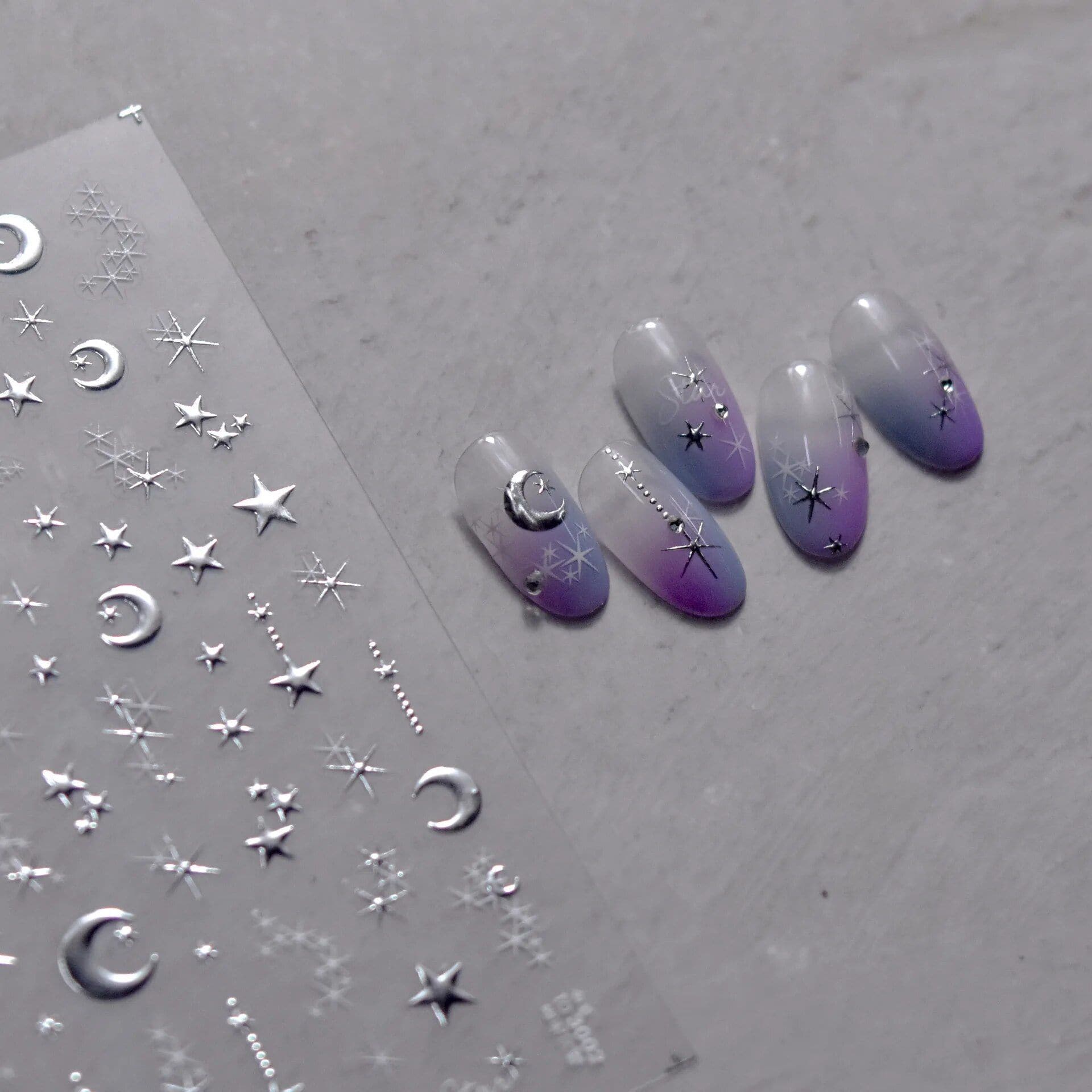 Elegant moon and star design nails with celestial motifs on a gradient purple and gray background. Perfect for stylish, themed nail art enthusiasts seeking unique nail decor.