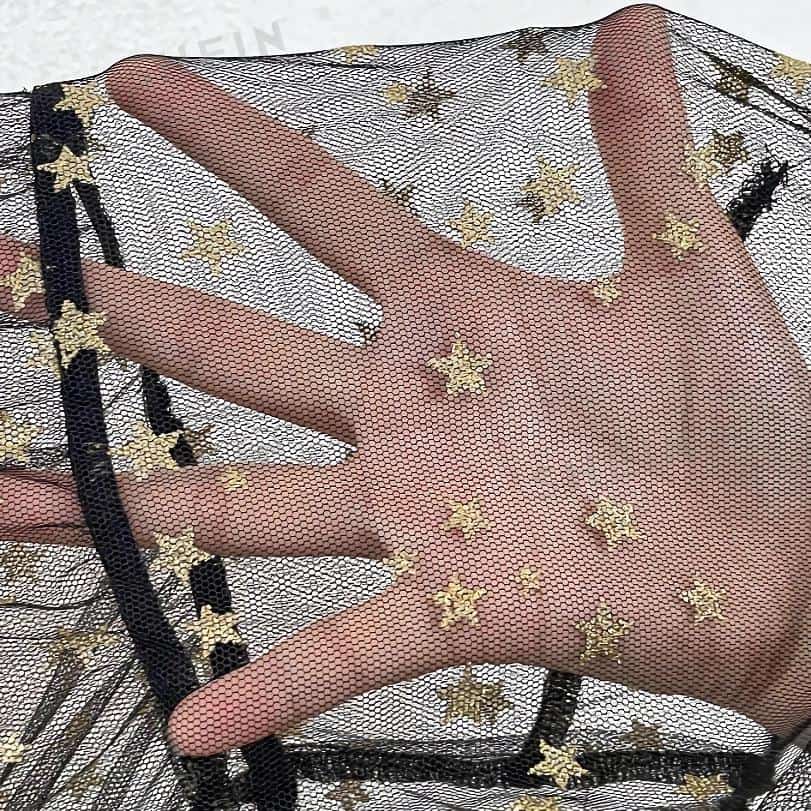 Close-up of a hand with black-painted nails wearing a black ring, against a sheer mesh fabric decorated with gold star patterns, promoting stylish accessories and jewelry at Monalisa Store.