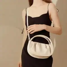 Elegant woman holding a stylish beige handbag with a modern design, wearing a black top with thin straps. Perfect for fashion-forward accessories and women's luxury handbag collections.