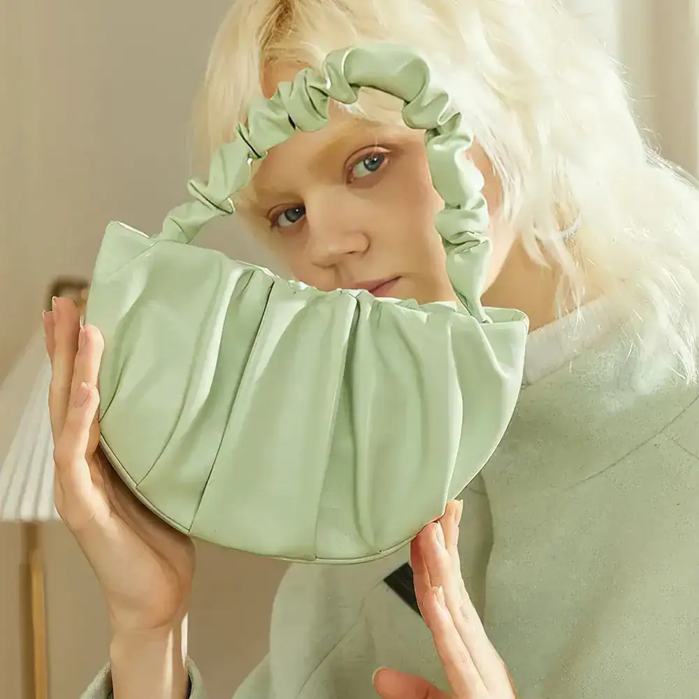 A woman holds a chic, pastel green handbag with ruched detailing, showcasing a modern, sustainable fashion accessory perfect for elevating your outfit.