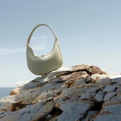 A stylish, off-white leather handbag with a curved design placed on a rocky surface against a clear sky, showcasing luxury fashion accessories.