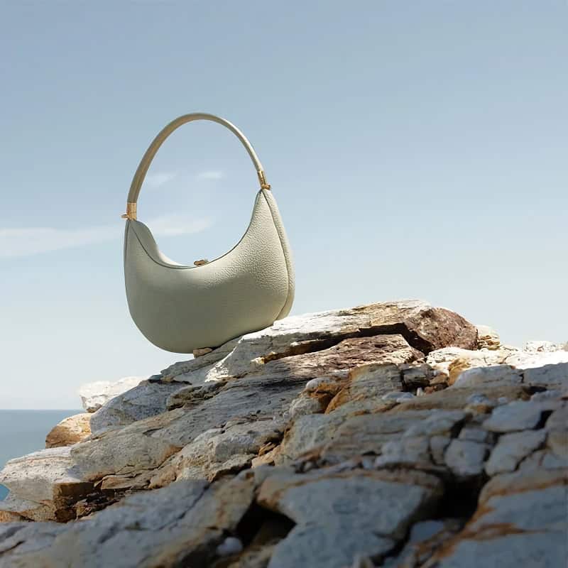 A stylish, off-white leather handbag with a curved design placed on a rocky surface against a clear sky, showcasing luxury fashion accessories.