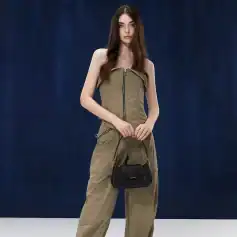 A stylish young woman with long, wavy hair wearing a strapless khaki jumpsuit holding a black designer handbag, standing confidently against a dark background.