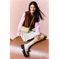 A fashionable young woman sitting on a brown surface against a pink background, showcasing trendy clothing, a yellow shoulder bag, and chunky black shoes, ideal for modern fashion and accessories from Monalisa Store.