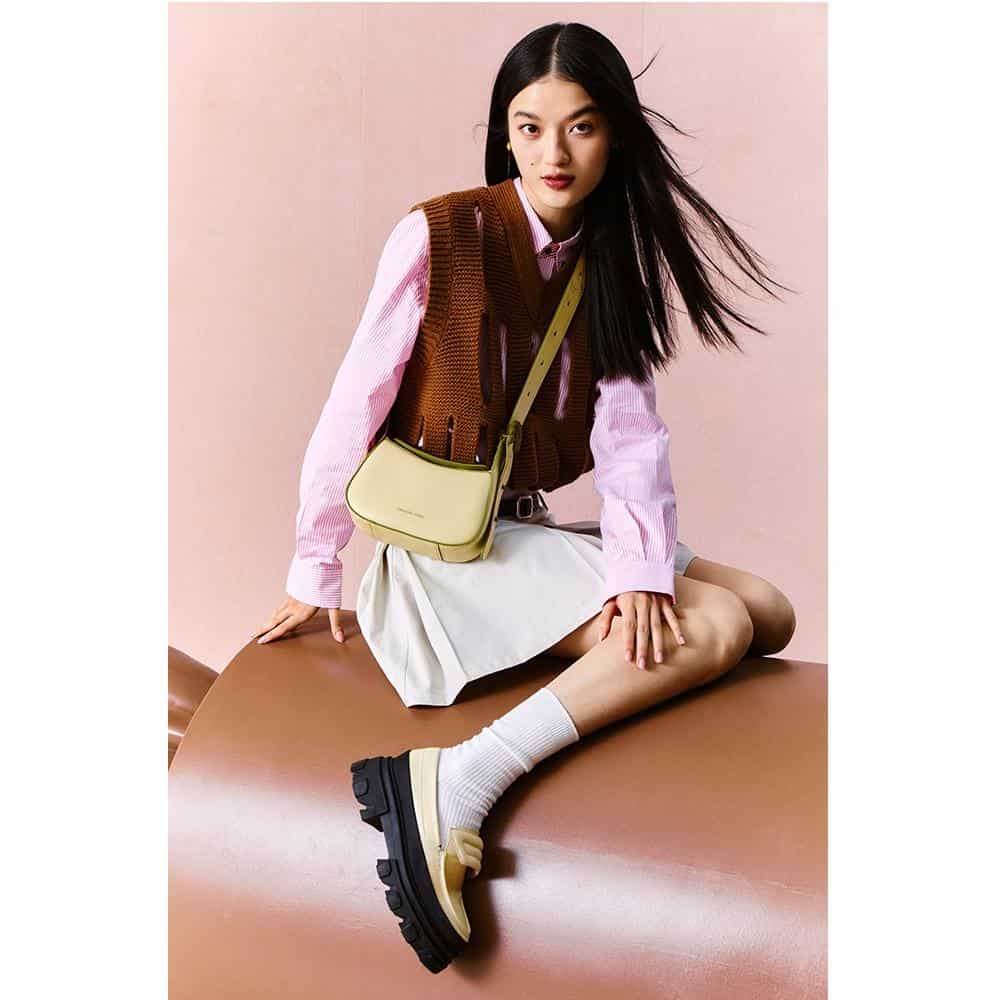 A fashionable young woman sitting on a brown surface against a pink background, showcasing trendy clothing, a yellow shoulder bag, and chunky black shoes, ideal for modern fashion and accessories from Monalisa Store.