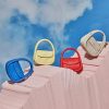 Vibrant handbags in blue, yellow, red, and beige displayed on pink textured surfaces against a blue sky backdrop.
