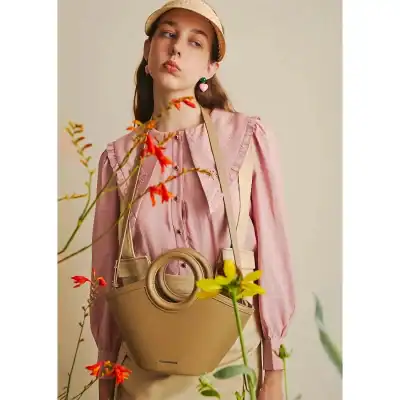Stylish beige handbag with circular handle, perfect accessory for trendy outfits. Featured in a fashion-forward look with pink blouse, capturing modern elegance.