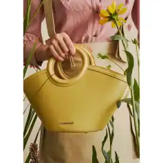 A stylish yellow handbag with unique circular handles held by a person wearing a pink shirt, surrounded by green plants and yellow flowers, showcasing trendy accessories for fashionable women.