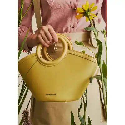 A stylish yellow handbag with unique circular handles held by a person wearing a pink shirt, surrounded by green plants and yellow flowers, showcasing trendy accessories for fashionable women.