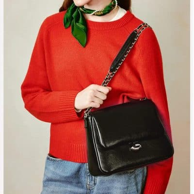 A woman holding a chic black leather handbag with a chain strap, dressed in a vibrant red sweater and casual jeans, showcasing elegant accessories available at Monalisa Store for fashion-forward customers.