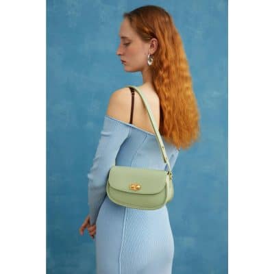 Stylish woman with long, wavy red hair, wearing a light blue off-shoulder outfit and earrings, holding a pastel green handbag, showcasing fashion accessories in an artistic photo.