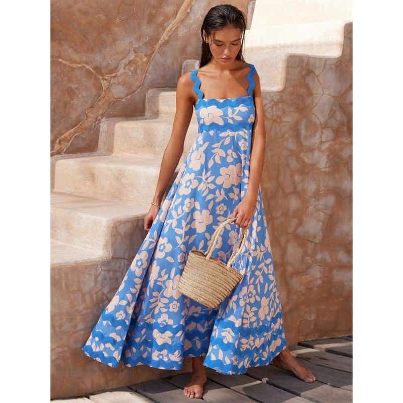 Beautiful woman in a blue and white floral maxi dress with ruffled straps, standing on a stairs with a woven straw handbag. Perfect for summer or vacation wear.