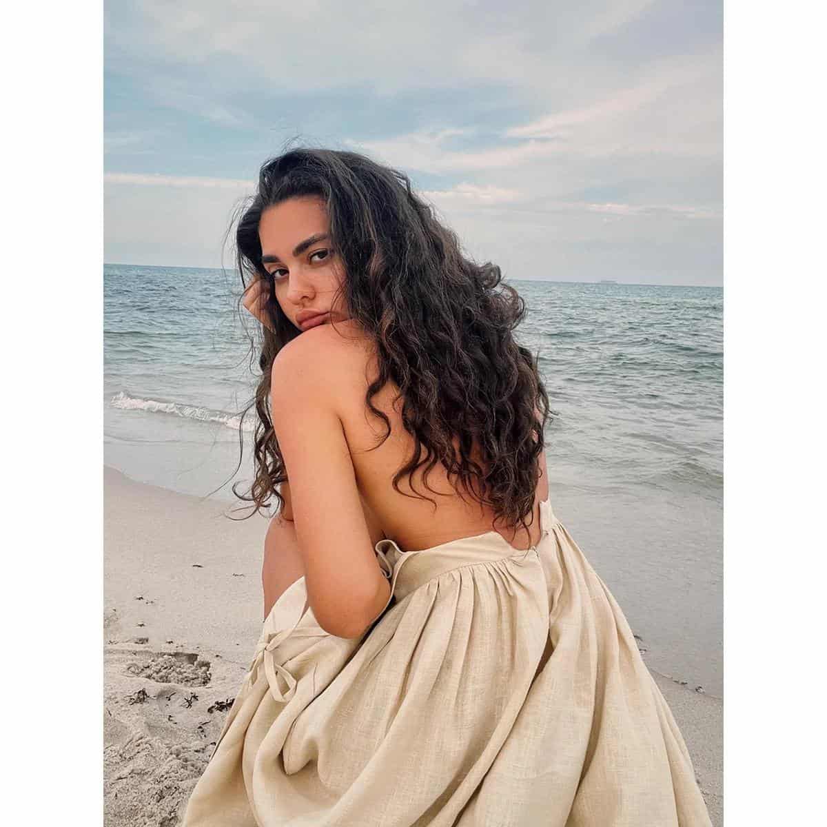 Close-up of a woman with curly hair sitting on a beach, showcasing stylish eyewear options available at Monalisa Store for optical needs.